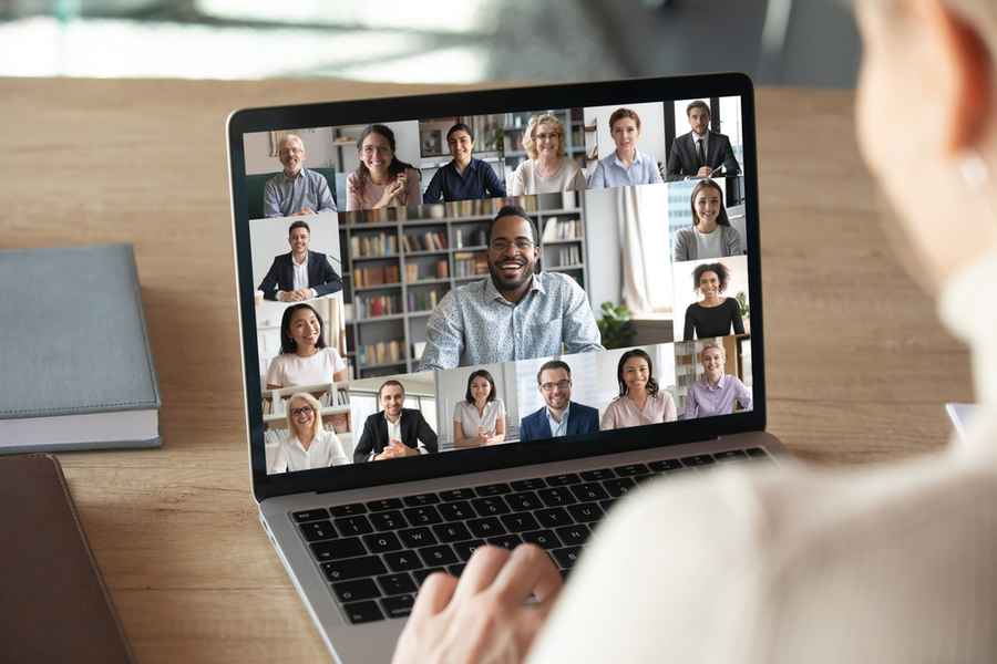 Photo of a person using a computer. On screen, a variety of smiling faces are revealed, similar to a video conference call.