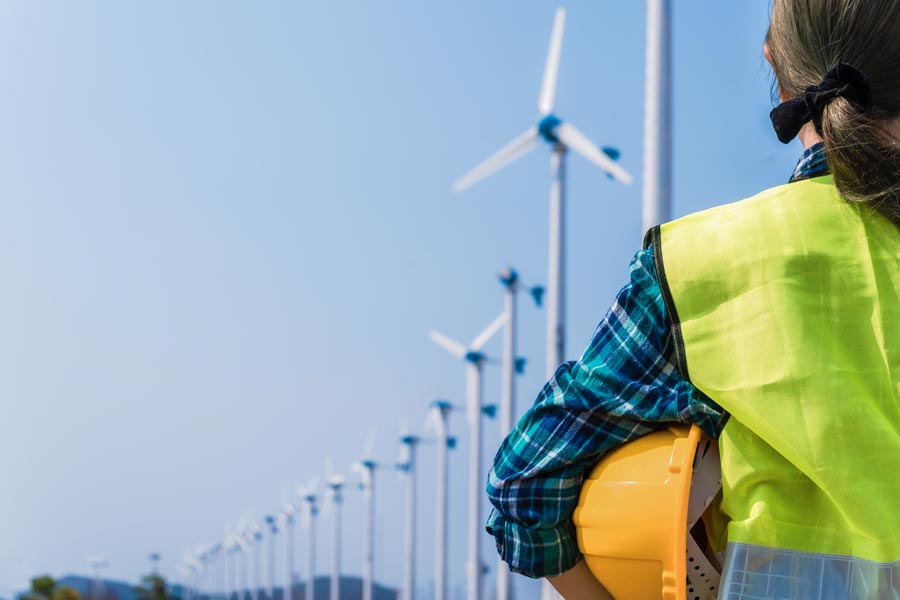 Woman holding a hard hat looks up at wind turbines in the distance.