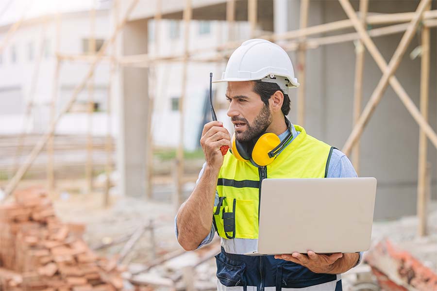 A male builder is talking into a walkie talkie while holding a laptop on a construction site.