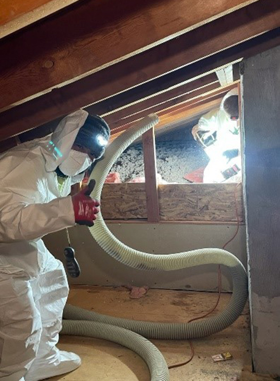 A construction worker in an attic blowing in insulation. The worker is wearing a protective suit and mask. 