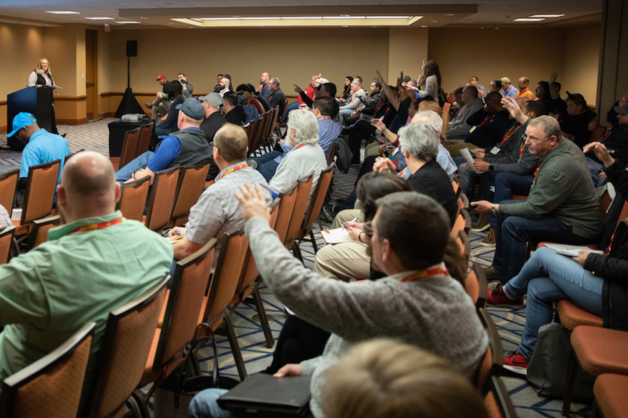 A group of people are in a conference room listening to a presenter. Several individuals have their hands raised.