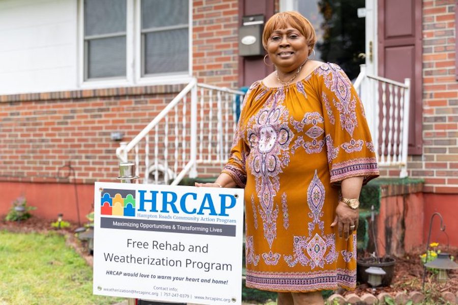 A woman standing in her front yard next to a sign that reads, "Free Rehab and Weatherization Program." She is smiling.