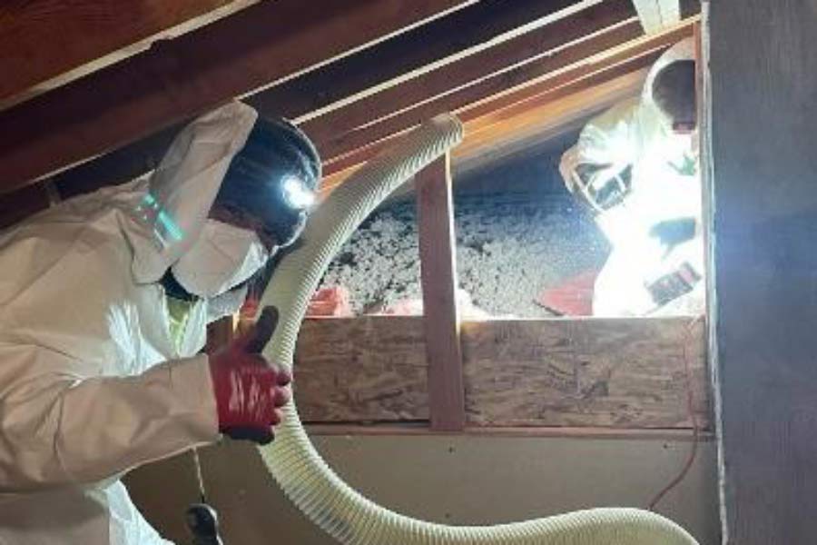 Two workers are in protective gear in an attic. They are using a device to add insulation. One person is giving a thumbs up sign on his hand.