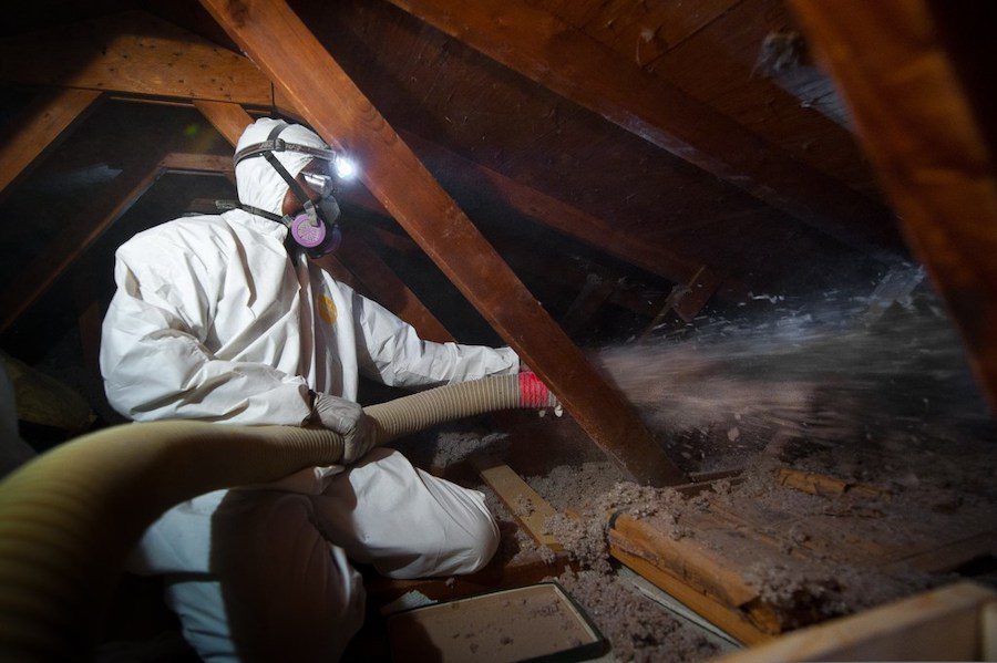 Man in attic in white personal protective suit blowing insulation onto the floor.