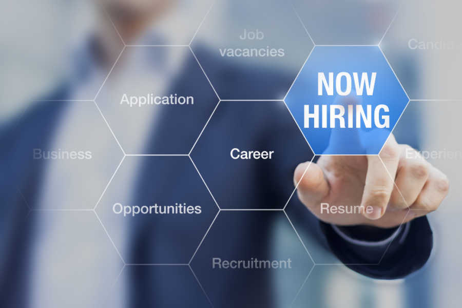 A man is blurry in the background of a graphic that is blue and shows him pointing to a sign that says now hiring.