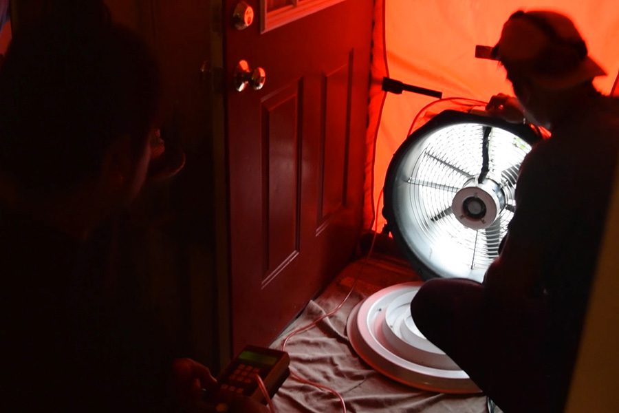 The sillouehette of a home performance contractor kneeling on the ground. The contractor is conducting a blower door test.