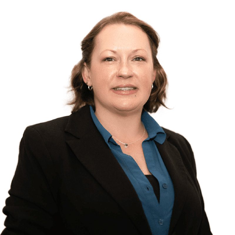 The image is of a woman standing on a plain white background. She is smiling and appears to be happy. The woman is wearing a black jacket and a blue blouse. The black jacket has a collar and is open at the front. The blue blouse has short sleeves and a round neckline. The woman's hair is not visible in the image, but she appears to have a natural skin tone. Overall, the woman looks relaxed and friendly.