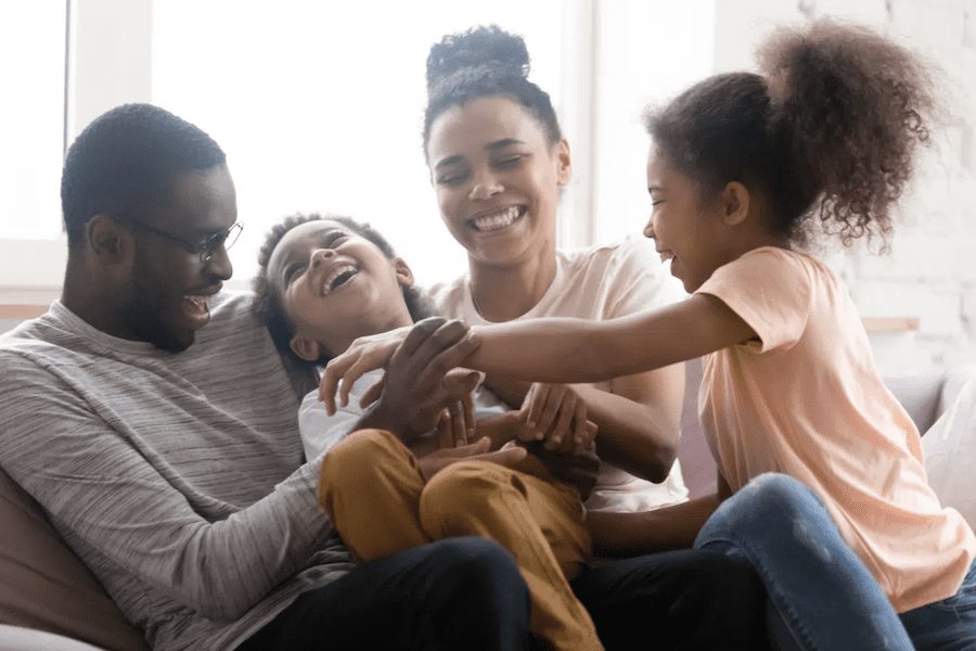 Family on sofa laughing together as sister tickles brother.