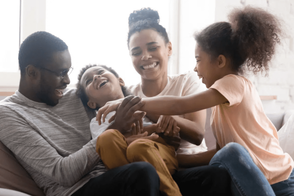 Family sitting on the sofa laughing while the sister tickles the brother.