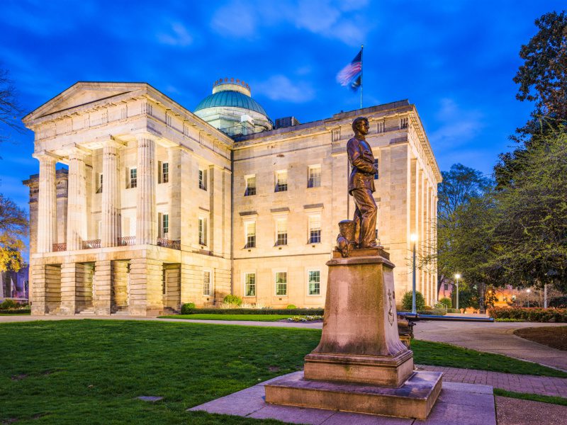 The North Carolina state capitol building with a bronze statue in front of it. The sky is dark blue and the building it lit up.