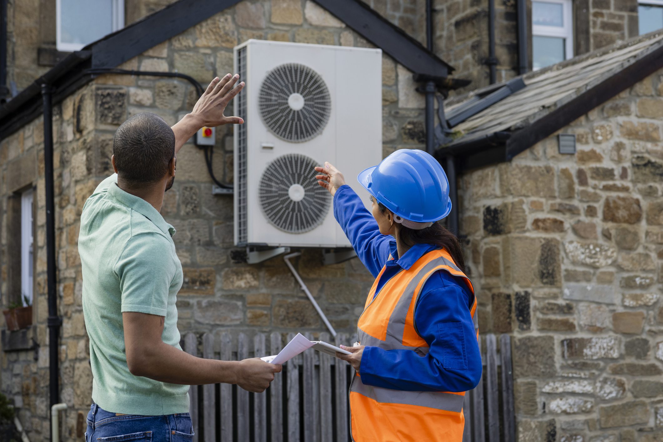An air source heat pump on the side of a home being installed in the North East of England. The house is aiming to be sustainable. There is a construction worker with a hard hat and reflective jacket holding a digital tablet, they are both pointing to the heat pump.